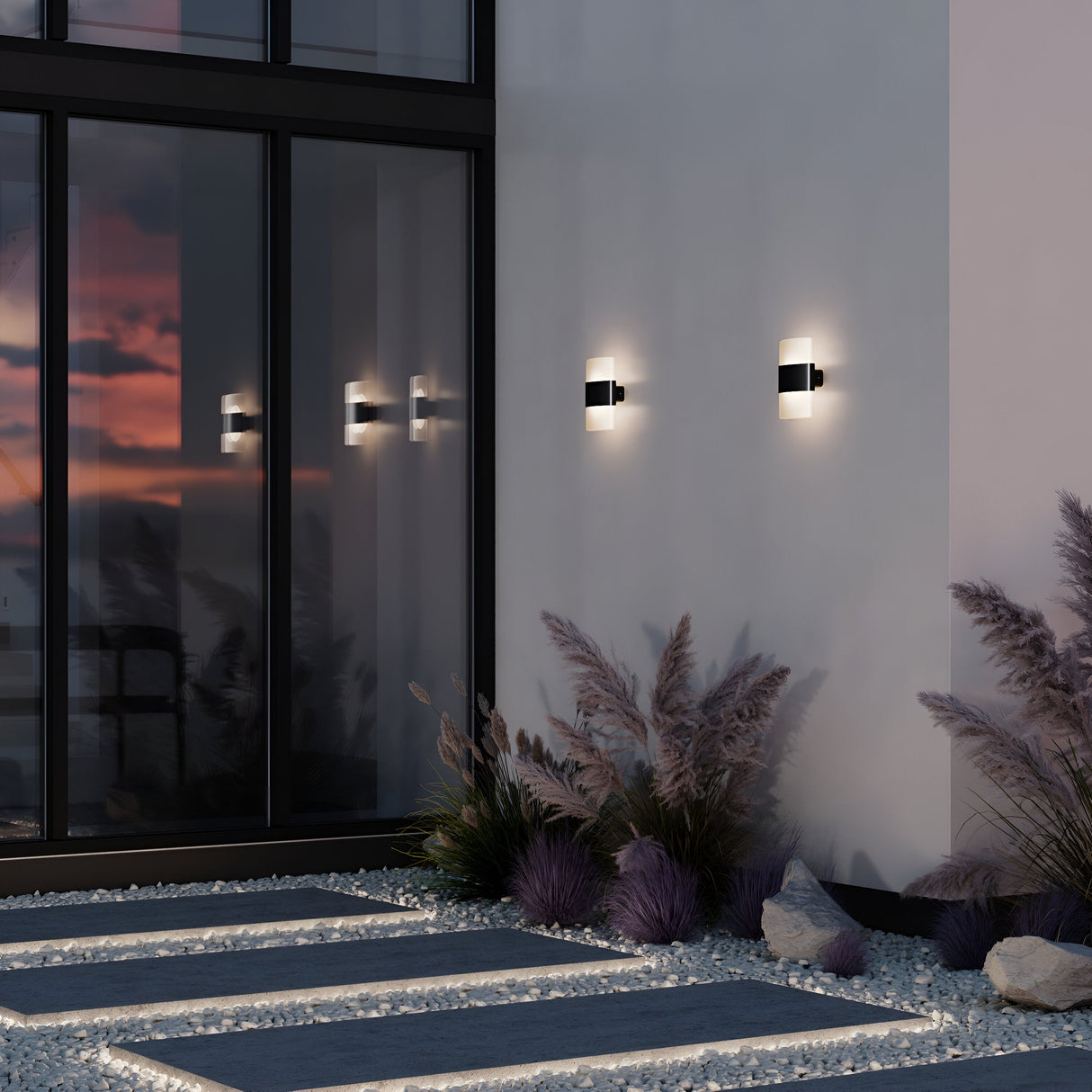 Rom Outdoor LED Up & Down Wall Lights illuminate a building exterior at dusk, their reflections visible in large glass doors. Ornamental plants and gravel decorate the ground below, with the Graphite finish anchoring the serene contemporary aesthetic.