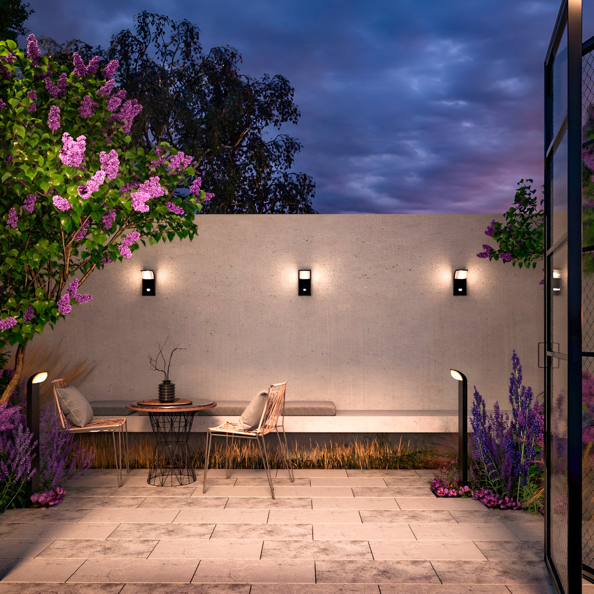 A cozy patio at dusk showcases a small table and two chairs under lilac trees. The concrete wall is adorned with the Stock Outdoor LED Wall Light 3000K 7W - Graphite, offering modern lighting. Lavender plants line the tiled floor, and a glass door is slightly ajar to the right.