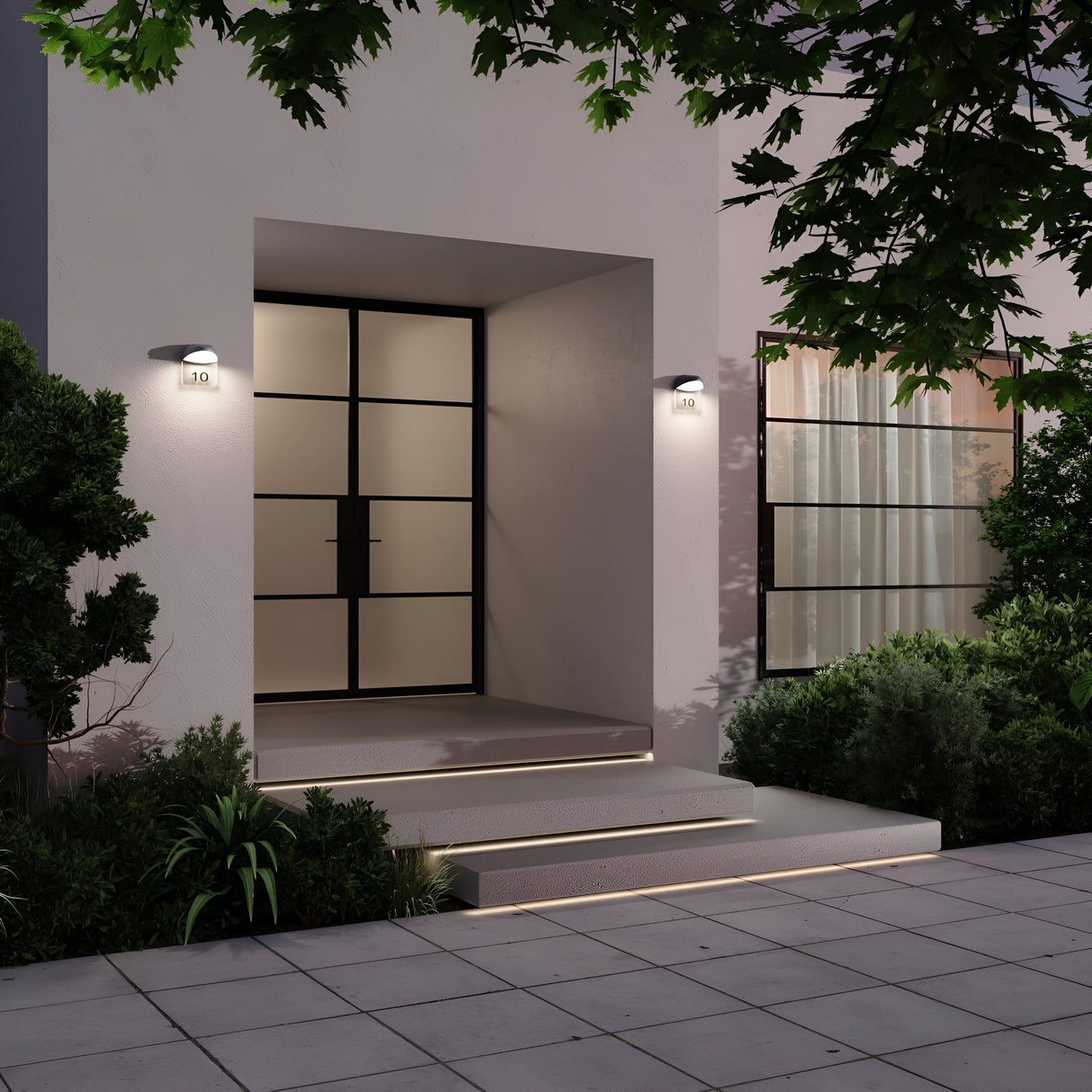 At night, a modern homes entrance showcases a large glass door with a black frame, brightened by two Gloss Outdoor LED Wall Lights in Graphite. These IP65-rated LEDs efficiently illuminate the steps leading to the door, which is bordered by lush green plants and trees.