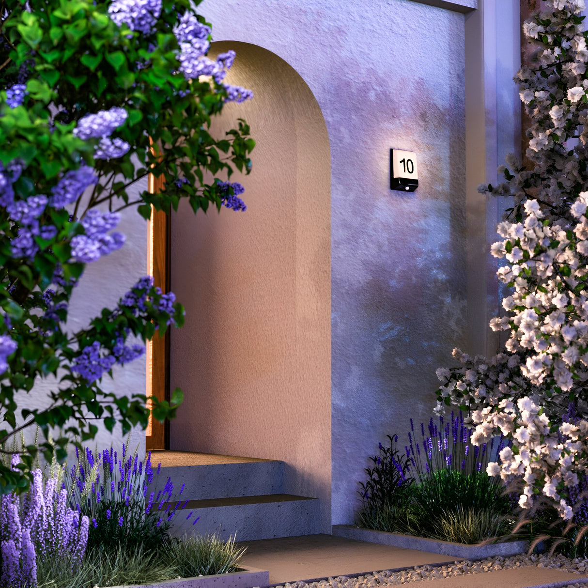 At dusk, a doorway marked 10 is illuminated by the Row LED Outdoor Wall Light 9W 3000K - Graphite, offering a serene welcome. Surrounded by lilacs and purple blooms, its IP44 rating ensures reliable protection.