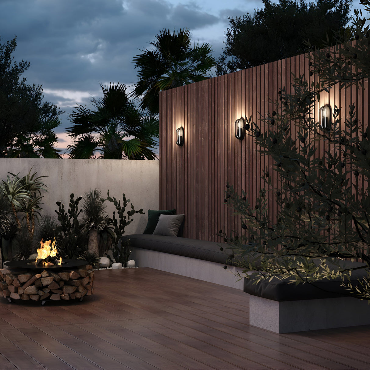 At dusk, a wooden wall on the rooftop patio features three Candle Outdoor Wall Lights in graphite. The space includes a cushioned built-in bench, surrounding greenery, and a fire pit with firewood on a wooden deck. Partly cloudy skies add to the contemporary loft-style ambiance.