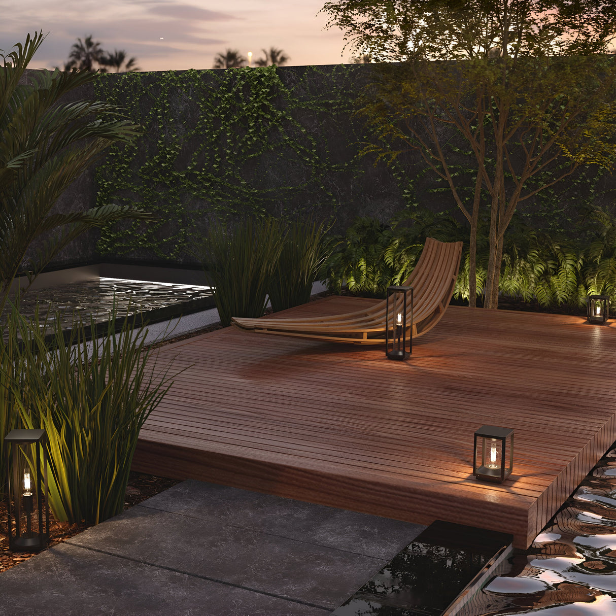 At dusk, a modern lounge chair and lanterns adorn the wooden garden deck. Cell Bollard Light 50cm in graphite finish illuminates lush greenery, tall plants, and a vine-covered wall. An outdoor water feature with floating stones enhances the tranquility of this serene area.