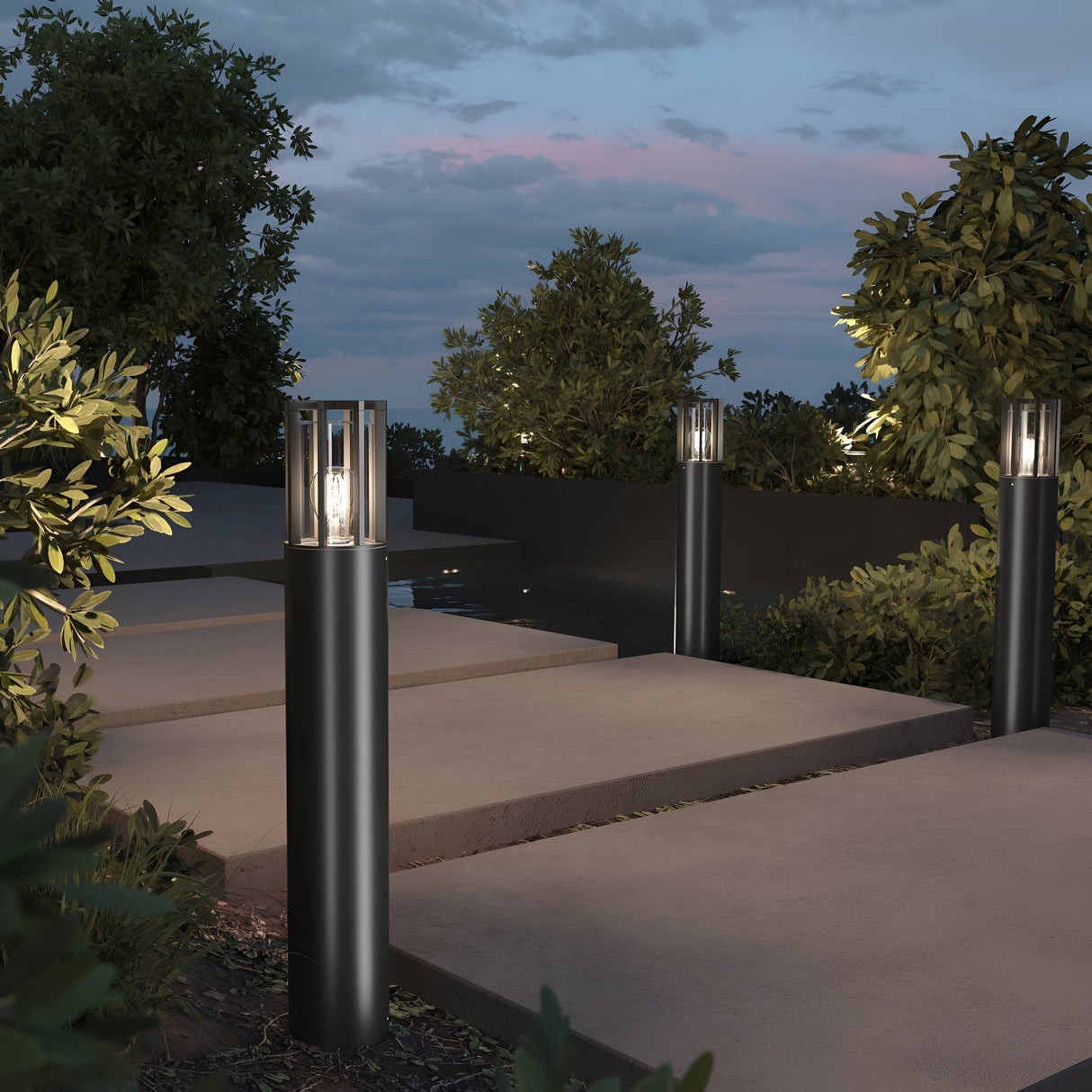 Elegant Barrel Bollard Light 75cm in graphite illuminates the path at dusk, casting a warm glow on concrete bordered by lush green bushes. Above, hints of sunset colors peek through partly cloudy skies.