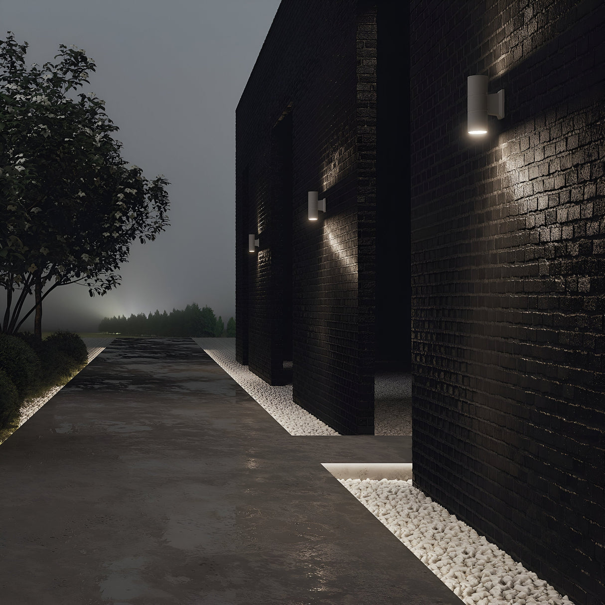 A dimly lit path with black brick walls is adorned with the Bowery Outdoor 2 Light Wall Light - Grey (O574WL-02GR). White pebbles line the base, and a tree stands along the path. The bidirectional light adds both style and safety to this mysterious, foggy setting where more trees emerge.