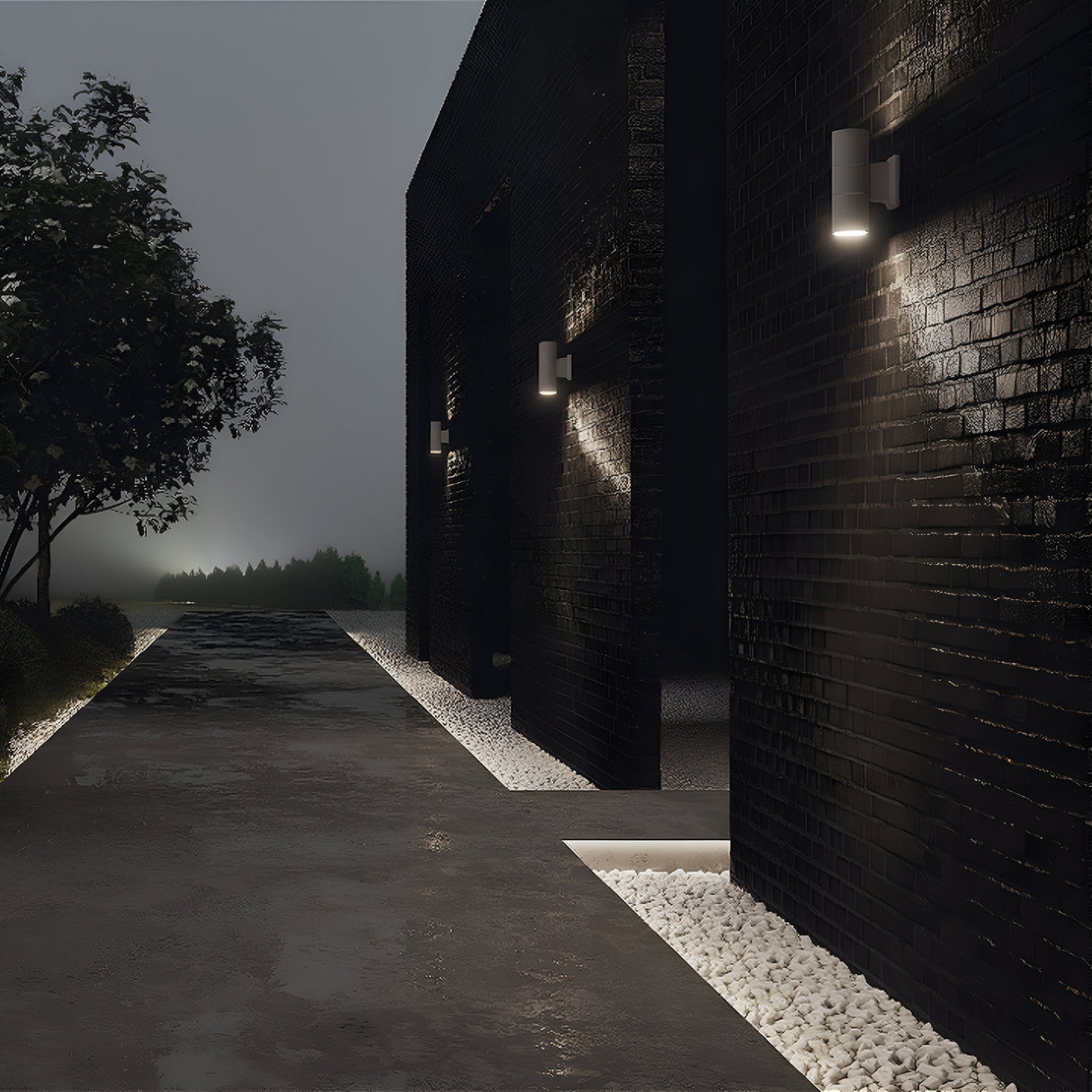 At dusk, a modern building with black brick walls is illuminated by Bowery Outdoor 2 Light Wall Lights in white (O574WL-02W) for dual light output. A pebble-lined path leads to a tree on the left under an overcast sky, highlighting the sleek elegance of contemporary architecture.