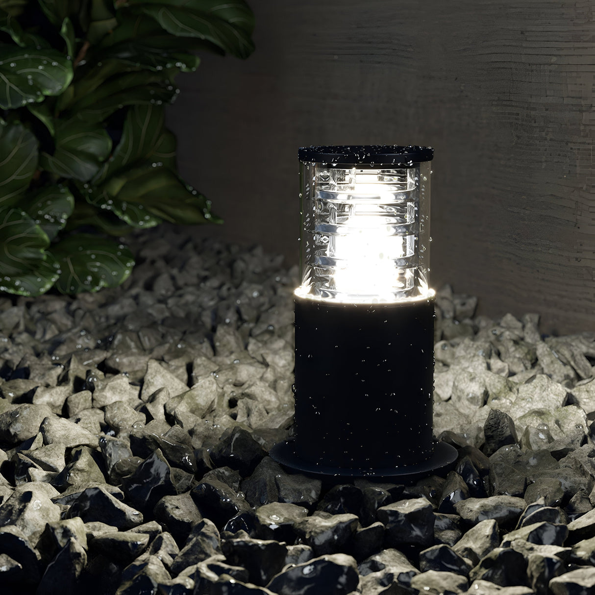 The Bronx Bollard Light 796mm - Black, crafted from corrosion-resistant aluminum, stands on wet, dark gray rocks near a leafy green bush. Water droplets cover the lantern and surroundings post-rain, softly illuminating the area and enhancing the charm of outdoor lighting.