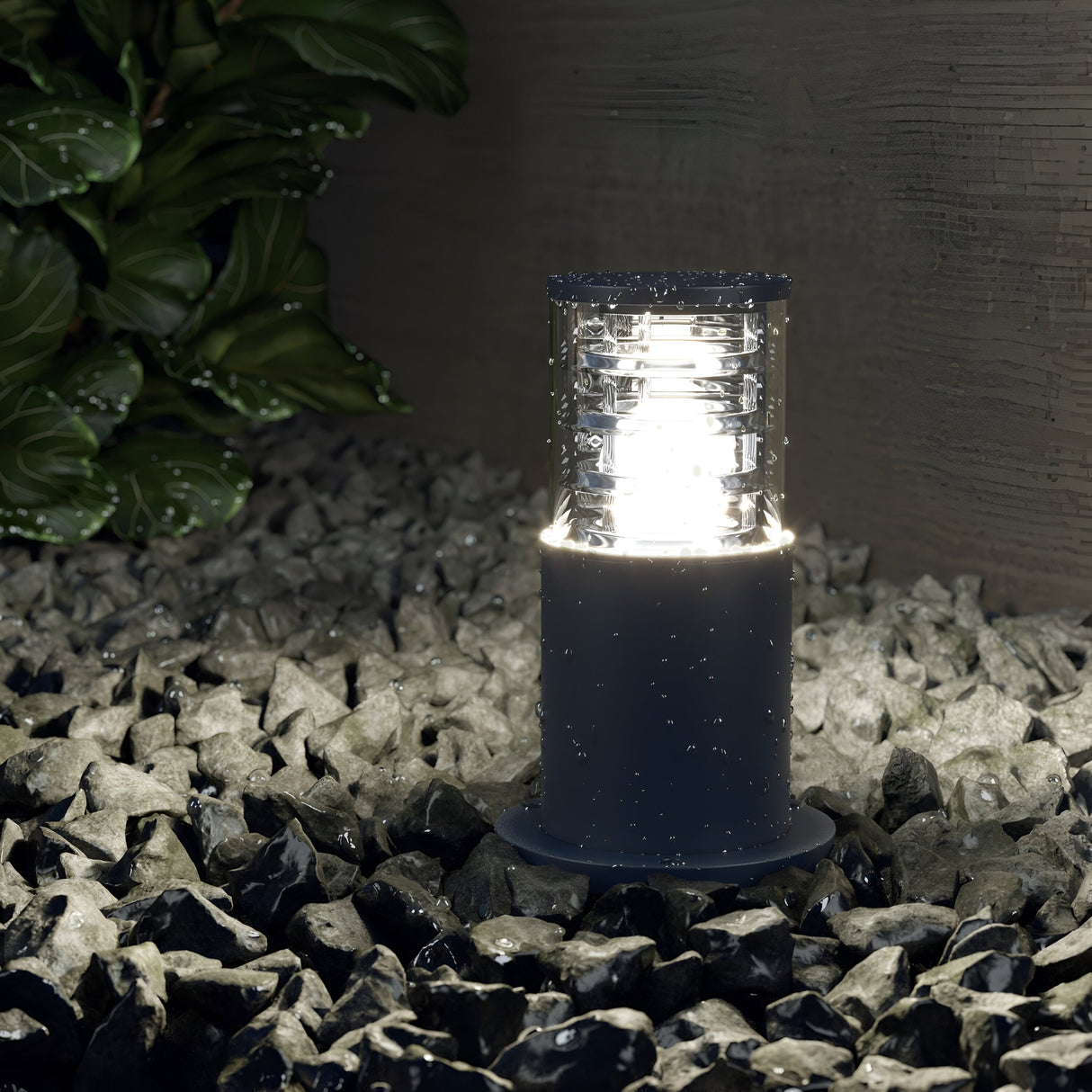 The Bronx Bollard Light 250mm - Grey stands on gray stones, casting a bright glow that illuminates glossy surfaces. Its corrosion-resistant design ensures durability, complemented by green foliage in the background for a natural touch.