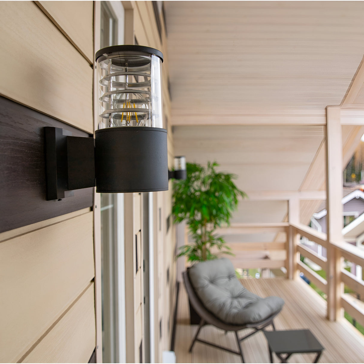 The Bronx Outdoor Wall Light - Black, with a modern cylindrical design, is mounted on a beige-paneled wall of a wooden balcony. In the background are a gray cushioned chair, a plant, wooden railings, and partial house views, offering both style and corrosion resistance.