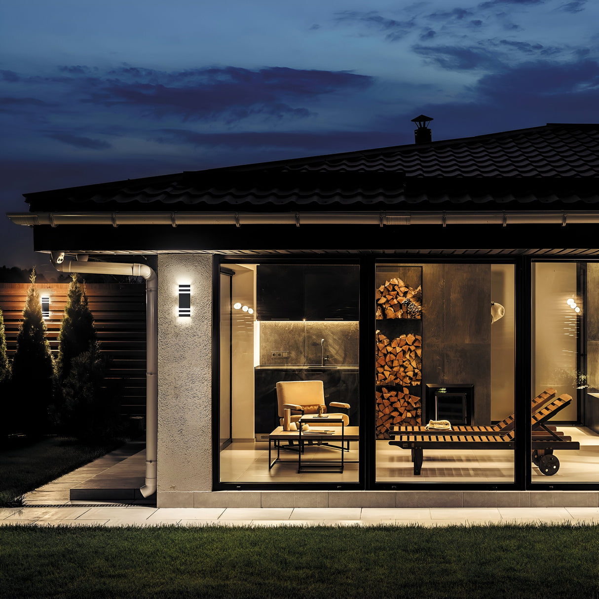 At dusk, a modern house features large glass windows with a warmly lit interior. Inside, theres a lounge chair and stacked firewood. Outside, the well-kept lawn is softly lit by the Unter den Linden Outdoor Wall Light - Black (IP54) against the deep blue sky.