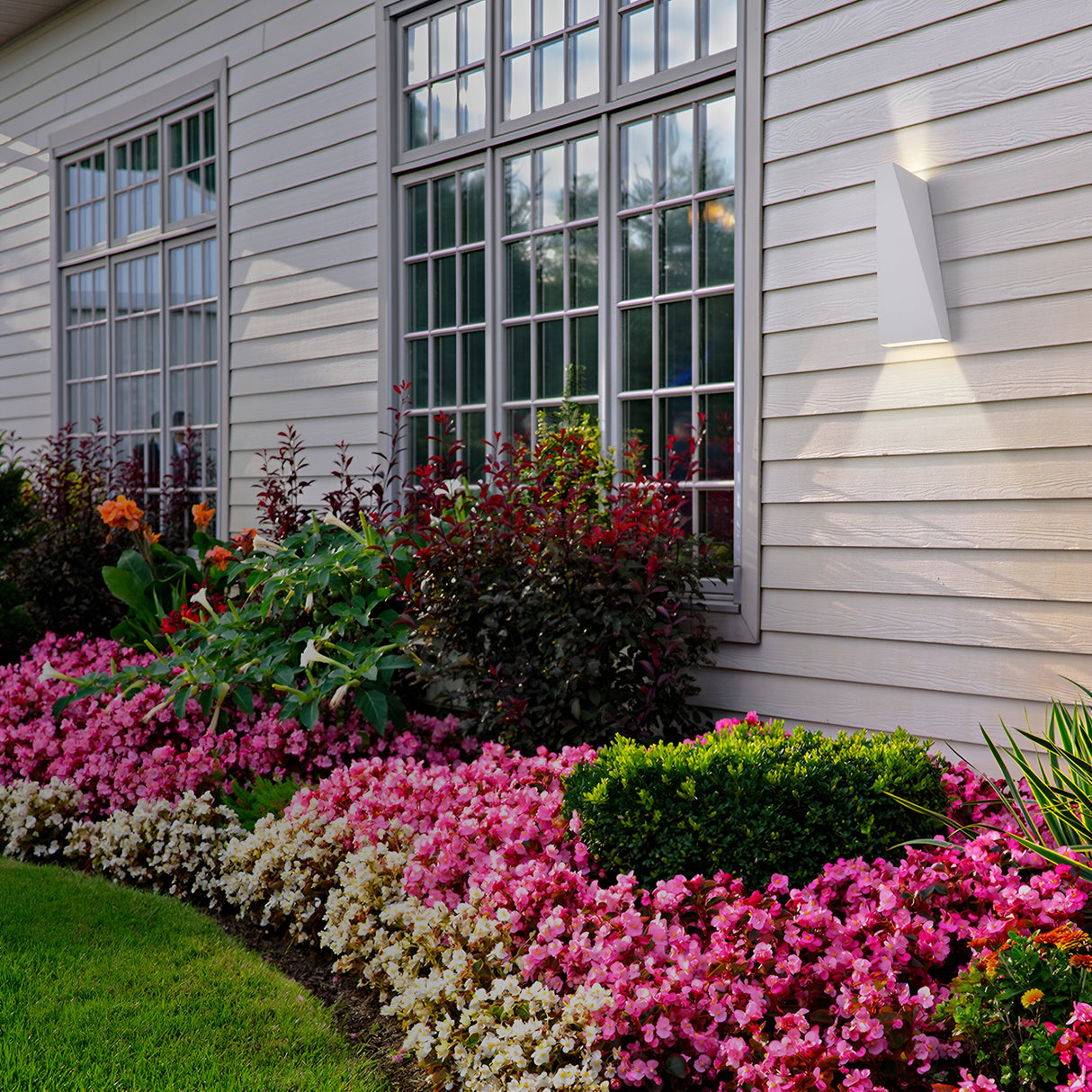 A house with white siding and large windows is highlighted by Times Square Outdoor Wall Light 3200K 6W - White, showcasing a well-maintained garden with thriving pink, white, and orange flowers alongside green shrubs on a neatly trimmed lawn.
