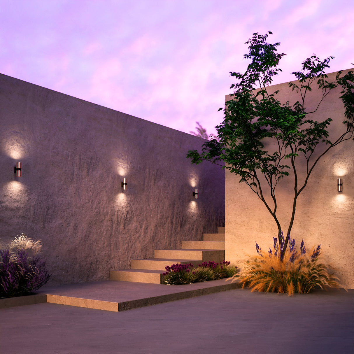 A serene courtyard with minimalist concrete walls and steps is softly lit by Bowery Outdoor 2 Light Wall Lights - Brass. A tree and ornamental grasses enhance the tranquility, while a pink and purple gradient sky adds to the ambiance with dual light output.