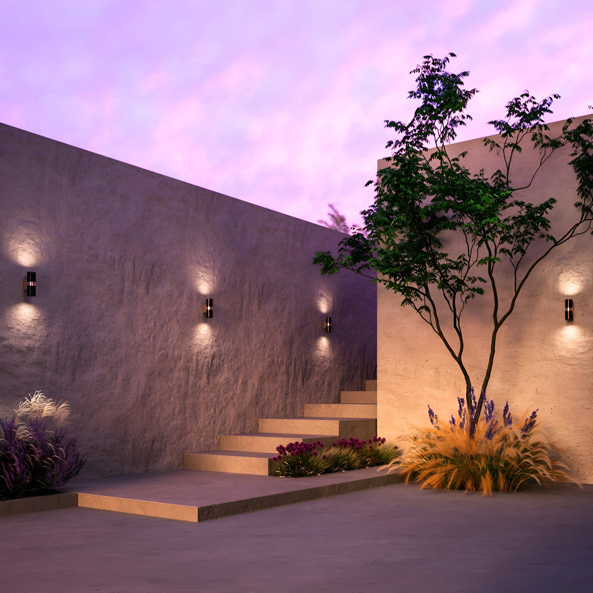 At dusk, a serene courtyard with minimalist stone walls and steps showcases the Bowery Outdoor 2 Light Wall Light - Black (O582WL-02B). Its brass accents and IP54 rating ensure durability, while soft lighting illuminates the scene, complemented by a tree and grasses against a vibrant purple-pink sky.