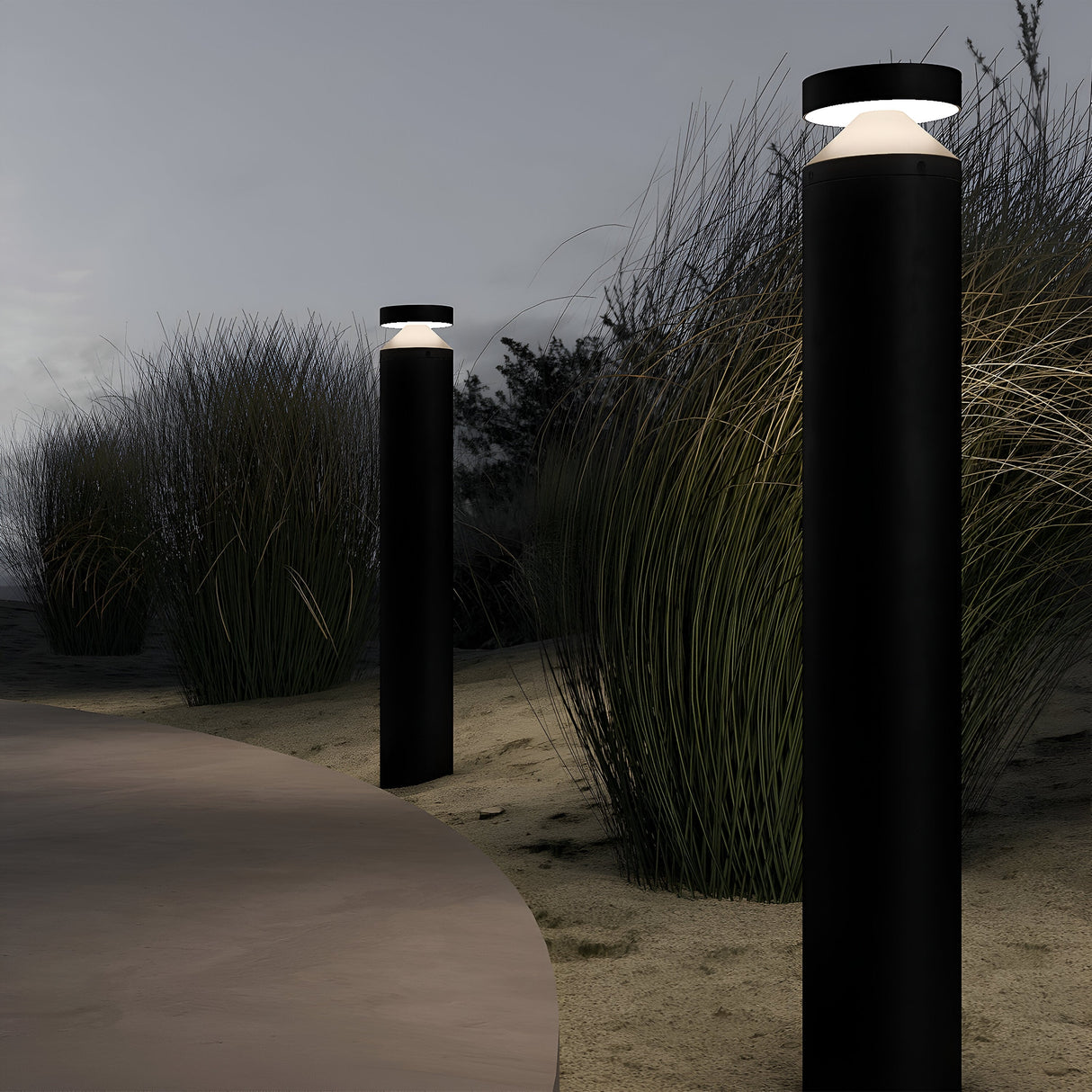 Two graphite Olten Bollard Lights, featuring an LED light source, illuminate a curved path bordered by tall grass under a dusky sky, enhancing the serene evening ambiance in outdoor spaces.
