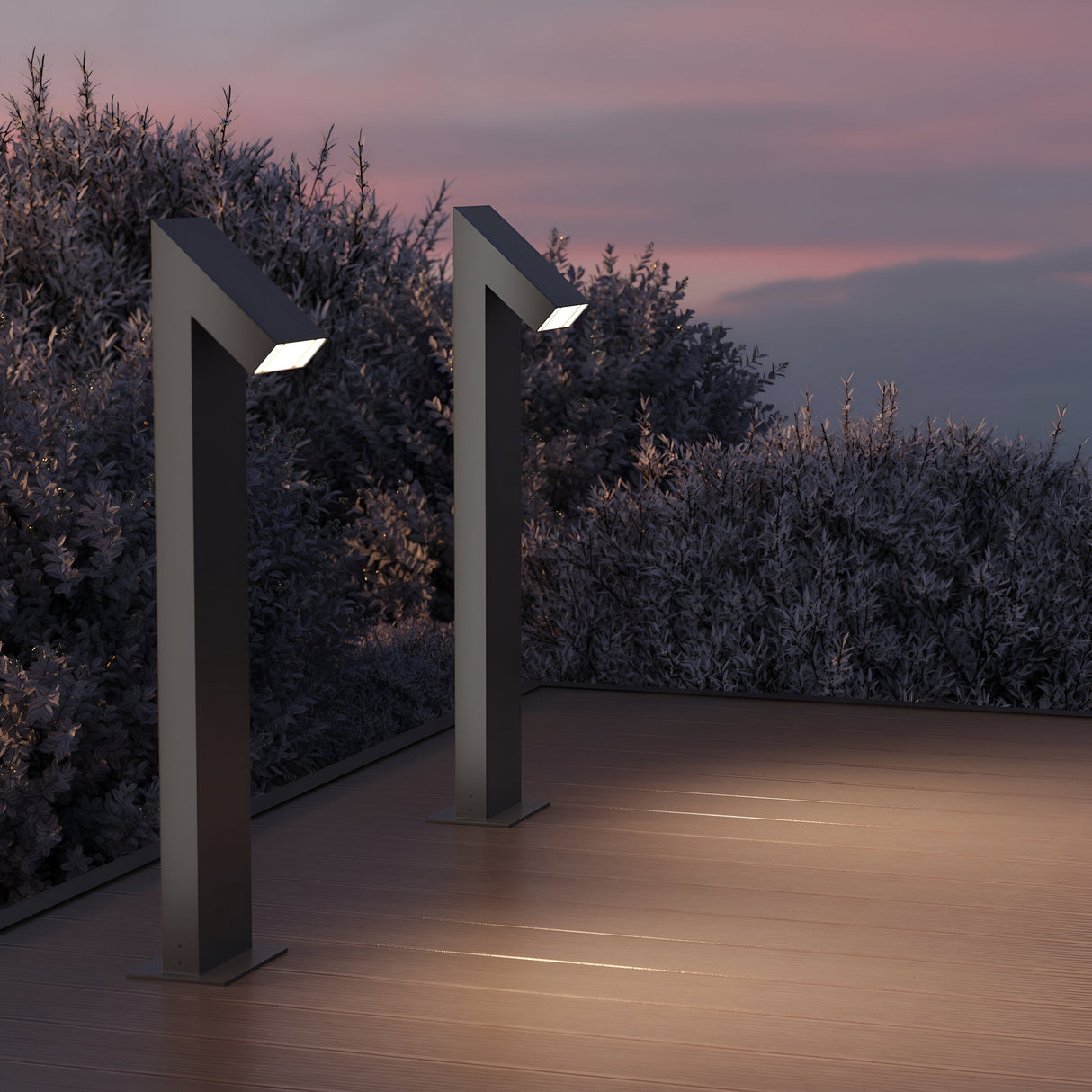 The Guss LED Bollard Light 90cm in Graphite lights up a wooden deck at dusk, with the sky in pink and blue hues and bushes around it adding tranquility. The IP65 rating of these fixtures ensures they withstand serene outdoor evenings with reliable LED illumination.