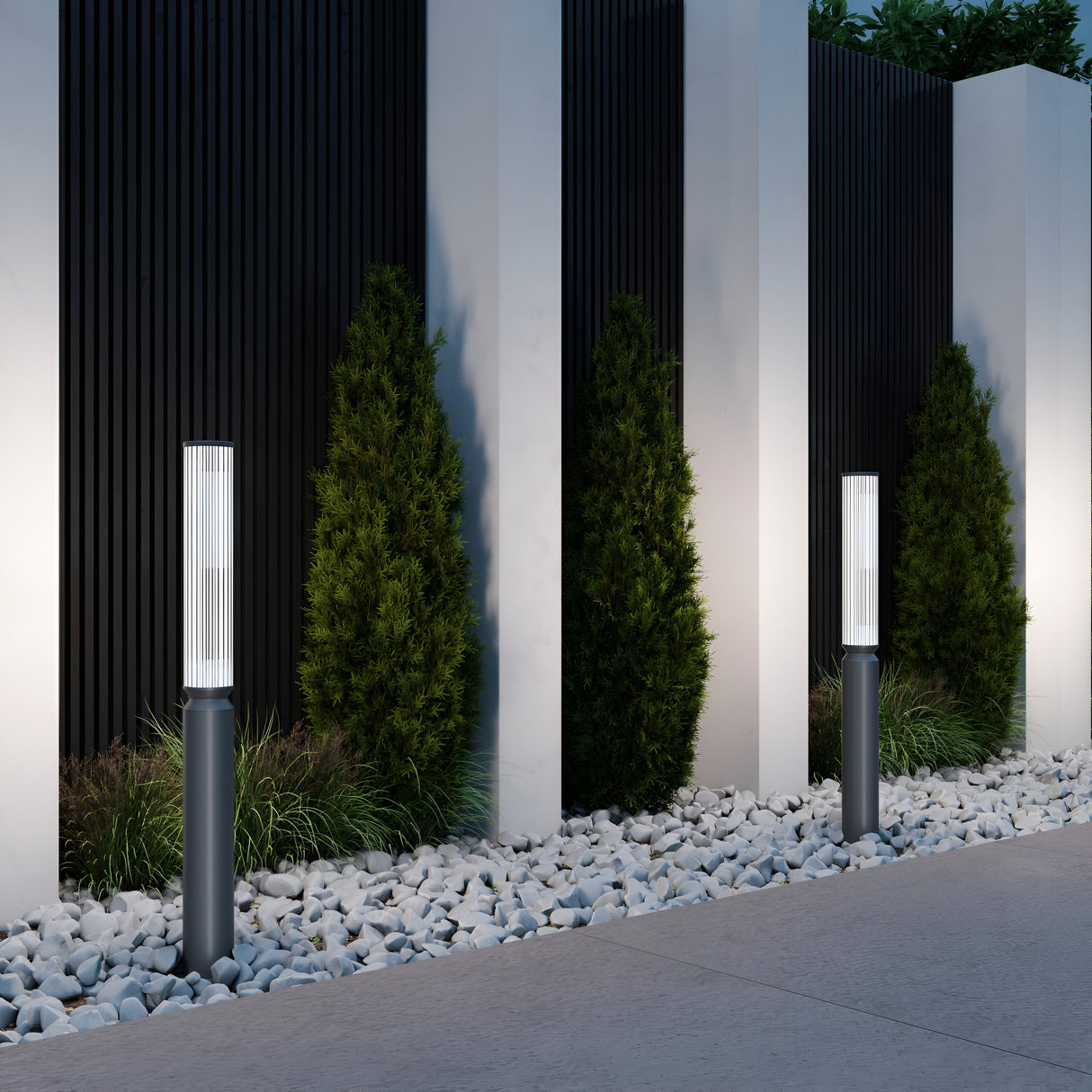 A modern garden with tall black metal posts featuring a graphite finish and vertical white panels. Small evergreen trees are evenly spaced, with white pebbles lining the walkway, enhanced by Lit LED Short Bollard Lights - Graphite, casting gentle shadows on the textured wall for a minimalist look.