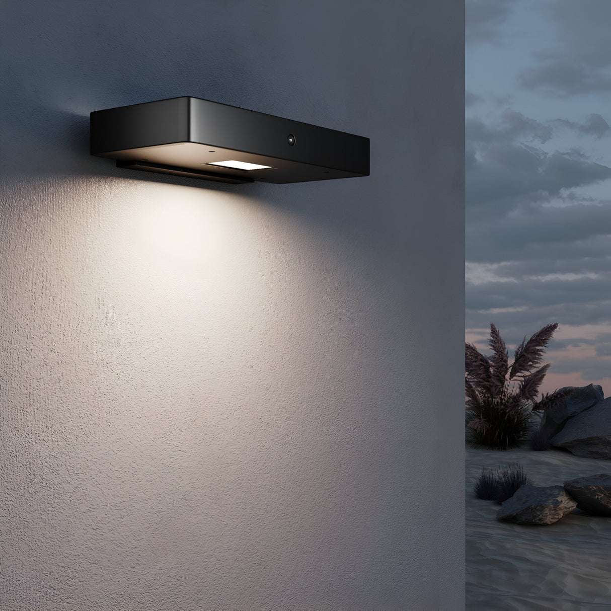 A Pir Outdoor LED Wall Light 3000K in Graphite casts warm light on a textured white wall, powered by solar energy. It enhances silhouettes of desert plants and rocks against a cloudy sky, suggesting an outdoor setting at dusk or dawn.