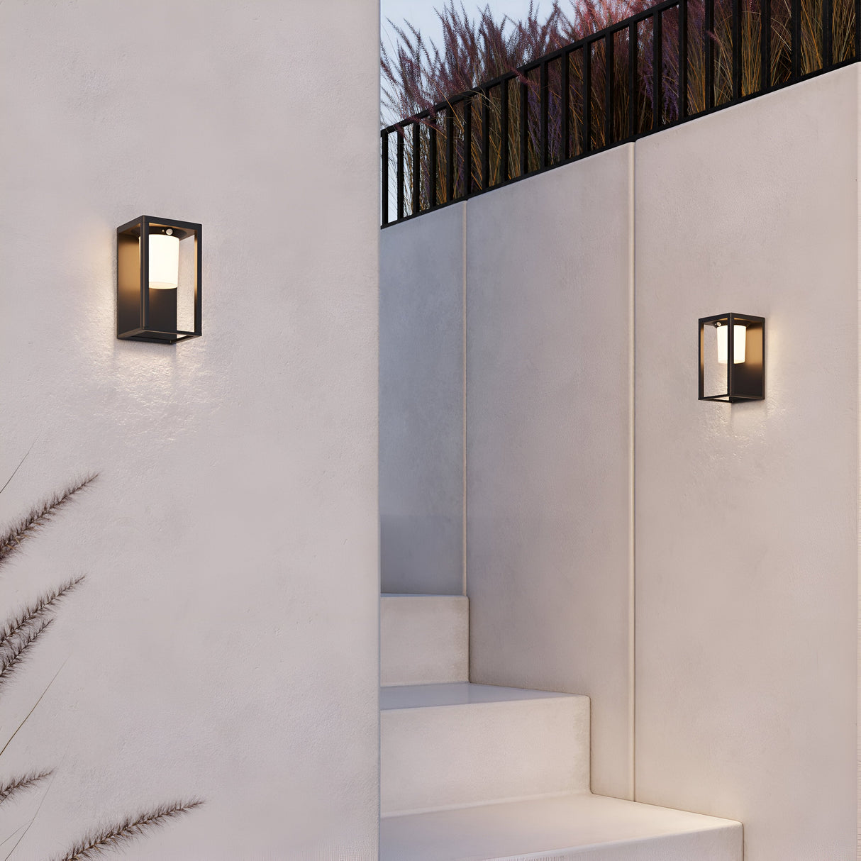 The Shatter Outdoor Wall Light 3000K - Graphite casts a soft glow on the minimalist white stairway, highlighting its geometric design. Equipped with solar batteries, they boost eco-friendliness. Tall grasses and a metal railing frame the scene at the top of the image.
