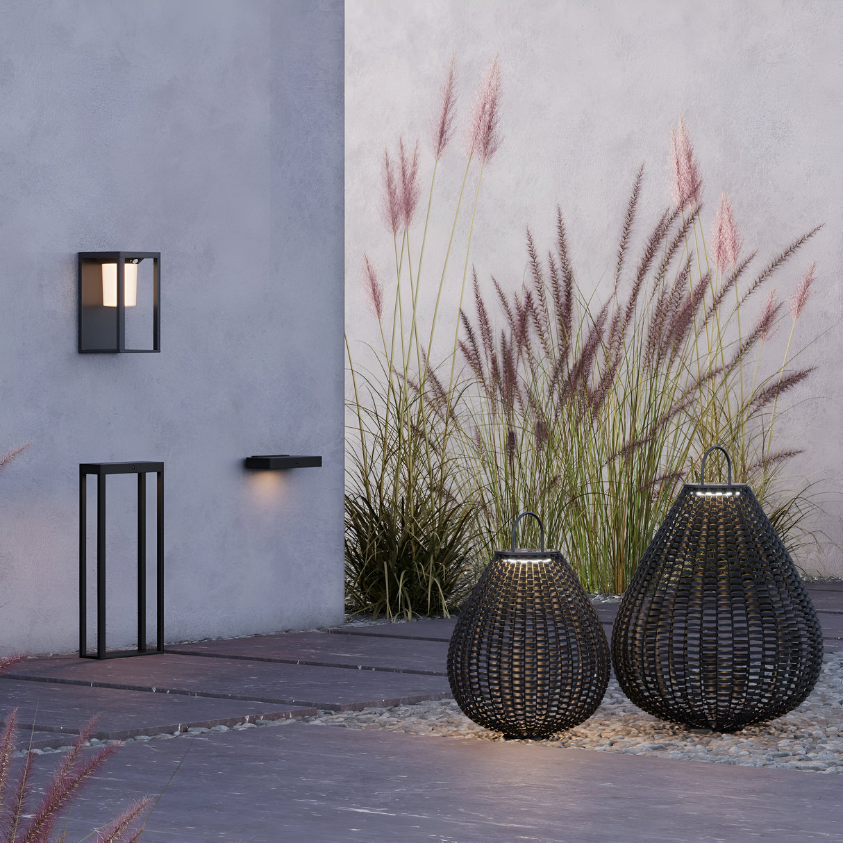 A minimalist outdoor setting includes two large black rattan lanterns on a stone patio, bordered by tall ornamental grasses. Outdoor lighting features a modern wall light and small black bench on a light gray wall, all efficiently powered by solar technology like the Plet Pedestal Light 61cm- Black.