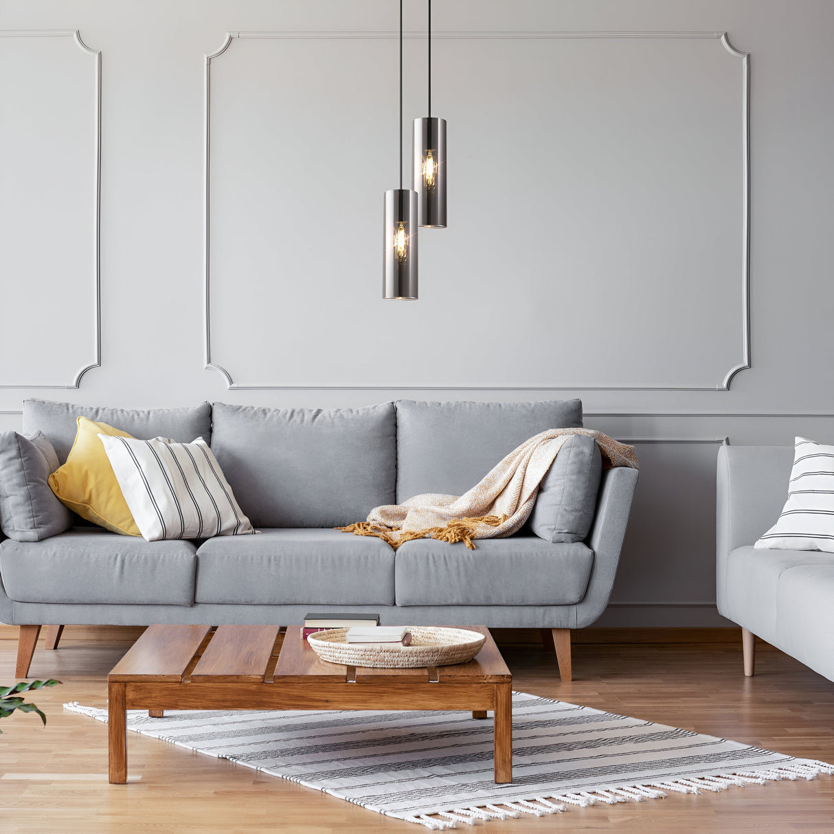 A modern living room highlights a gray sofa with striped and yellow pillows, accented by a beige throw. A wooden coffee table with a woven basket sits before it, under the Gioia Pendant Light - Chrome With Cylindrical Glass. A striped rug enhances the sleek aesthetic on the wooden floor.