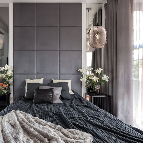A modern bedroom showcases a large gray upholstered headboard and a neatly made bed with black bedding and several pillows. Floral arrangements sit on two bedside tables, while the Dunas Oblong Pendant Light - Black with Smoked Patterned Glass enhances the contemporary aesthetic, casting soft light.