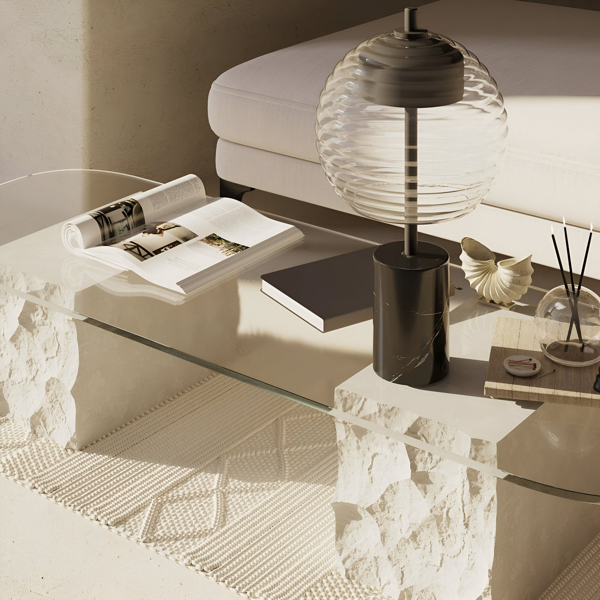 A modern glass coffee table with stone supports features an open book, a closed black book, the Mystic LED Table Lamp - Black With Marble Base, a seashell, and a reed diffuser. A cream sofa rests on a woven rug in the background, creating a cozy, minimalistic living space.