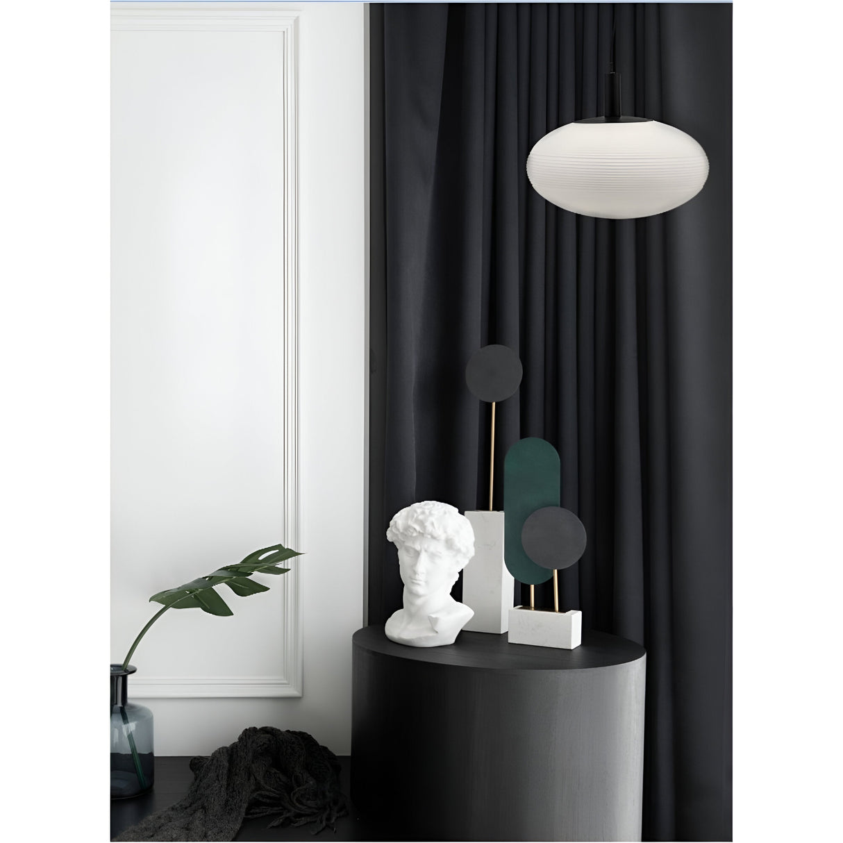 A minimalist interior showcases a white bust on a black pedestal, abstract sculptures on white bases, dark curtains, and the Perlas Pendant Light - Black With White Round Glass illuminating the space. A leafy branch in a glass vase completes this modern and artistic ambiance.