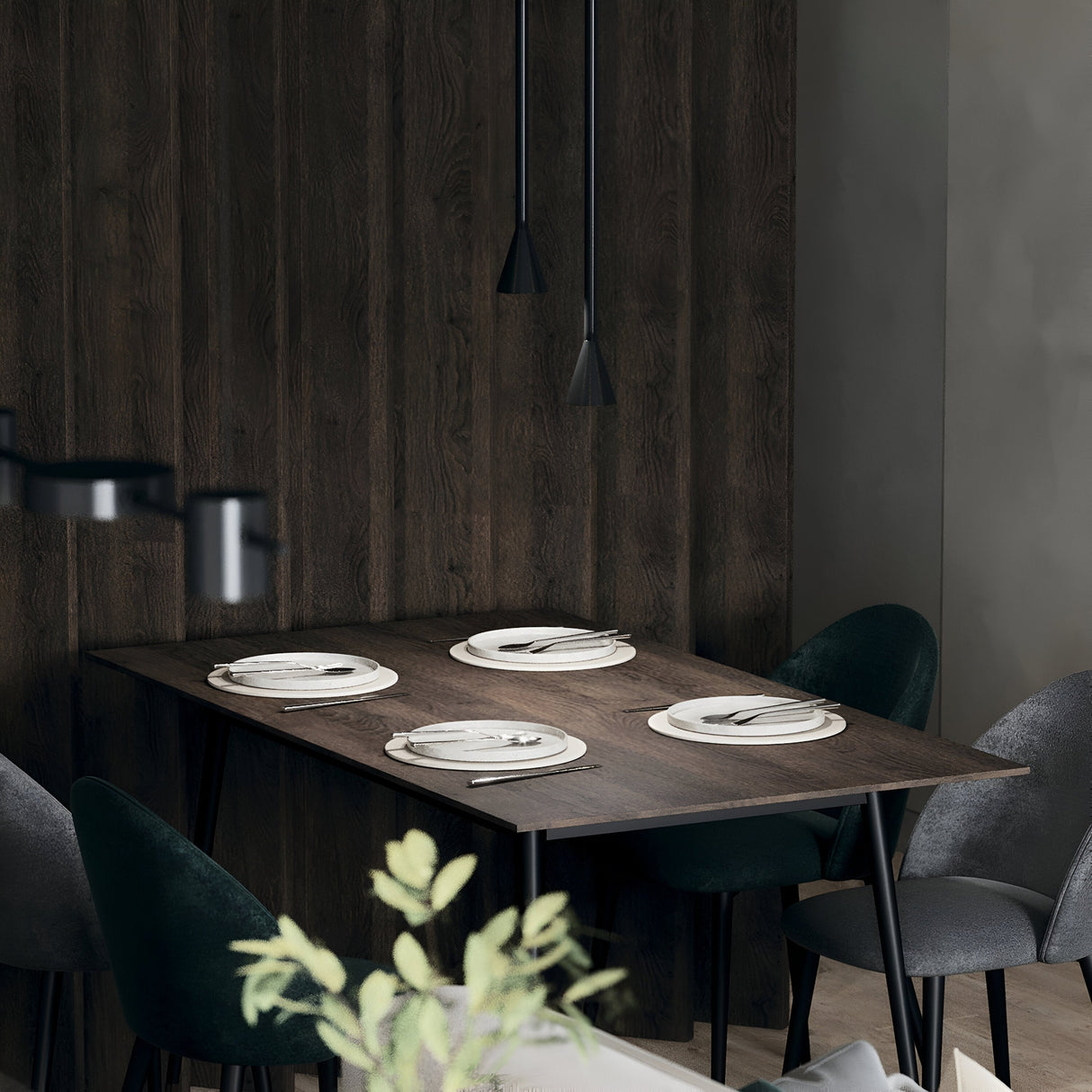 A modern dining area features a dark wooden table with four place settings, surrounded by dark green chairs. The space is illuminated by the Arrow 1 Light Long Cone Pendant in black, and a blurred plant adds natural warmth in the foreground.