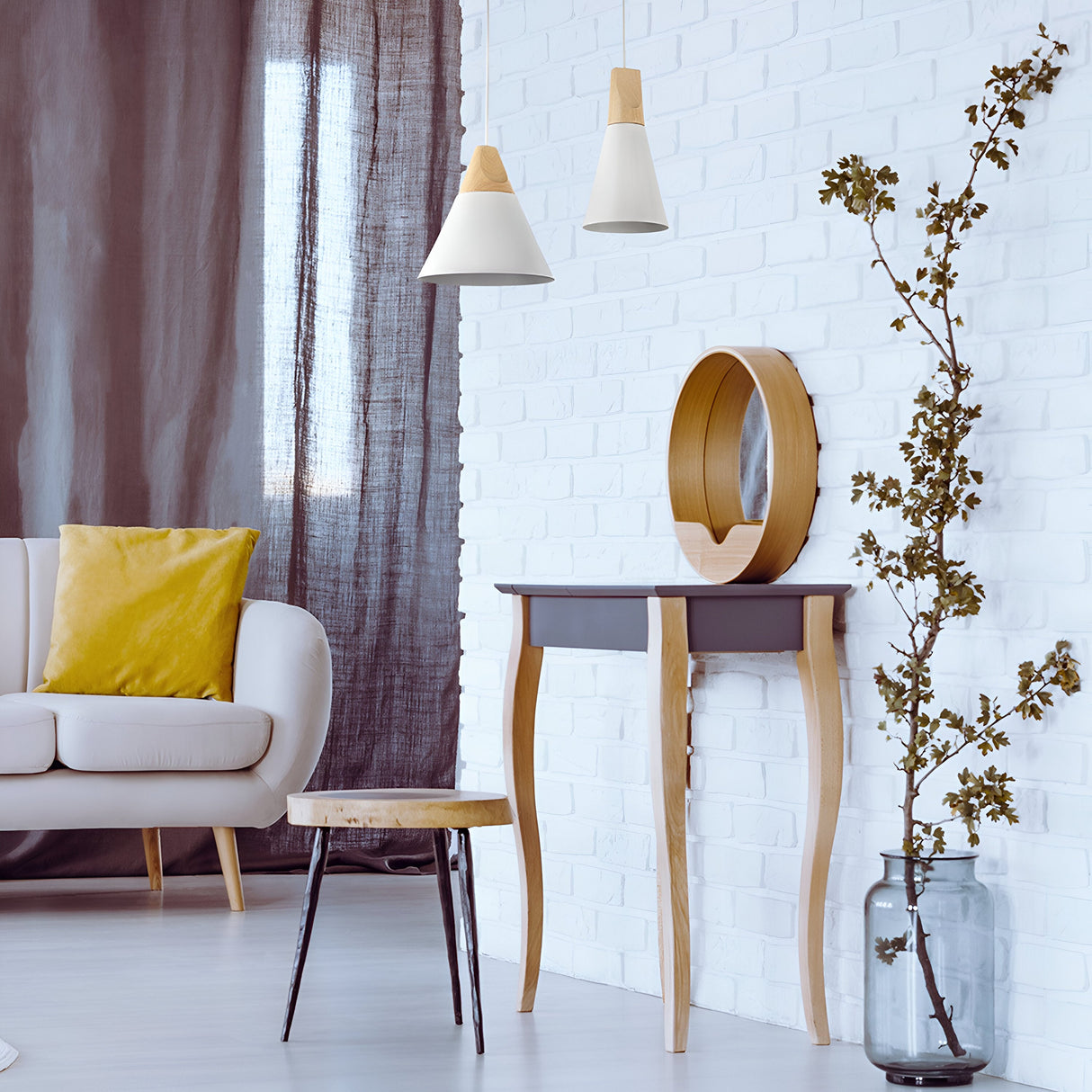 A modern living room showcases a white brick wall with a round mirror and a small curved-leg table. Pendant lights, including the 22cm Bicones Pendant Light in white with wood detail, hang overhead. A glass vase of branches adds natural elements, while a sofa with a yellow pillow sits on the left.