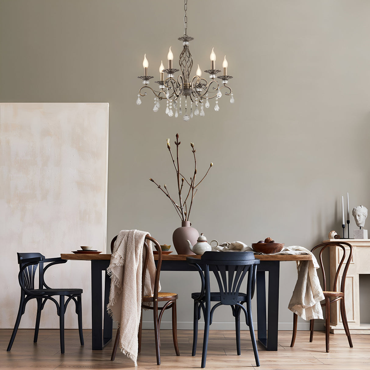 A dining room showcases a wooden table with assorted chairs adorned with vases and branches, while a plush throw rests on one chair. The Grace 6 Light Chandelier - Bronze hangs from the ceiling, featuring crystal pendants. A neutral wall serves as the backdrop for a sideboard holding decor.