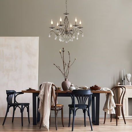 A dining room showcases a wooden table with assorted chairs adorned with vases and branches, while a plush throw rests on one chair. The Grace 6 Light Chandelier - Bronze hangs from the ceiling, featuring crystal pendants. A neutral wall serves as the backdrop for a sideboard holding decor.