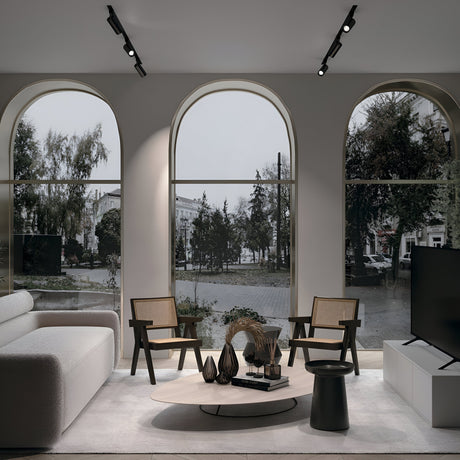 A modern living room with large arched windows overlooks a tree-lined street. It features a white sofa, wooden chairs, and a round coffee table adorned with decorative vases. The sleek TV on a stand is enhanced by the Focus Single Phase Unity Track Spotlight in black for visual comfort.