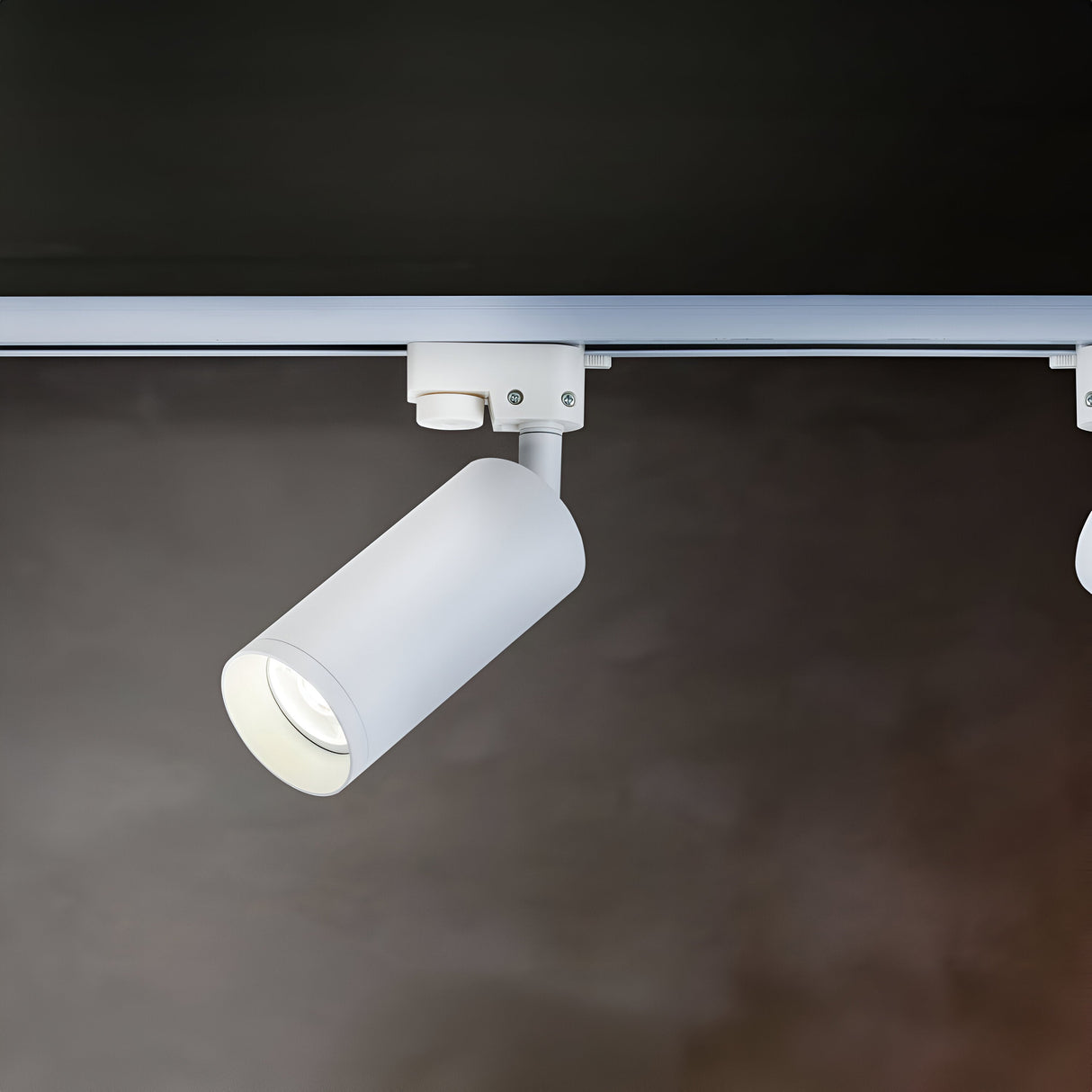 The Focus Single Phase Unity Track Spotlight - White, with a cylindrical design and GU10 bulb compatibility, is mounted on a straight rail against a dark textured background. It features a swivel mechanism for downward illumination of the space below.
