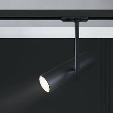 The Elti Single Phase Unity Track Spotlight - Black features a sleek, modern cylindrical design, hanging from a single-phase track system. It casts a focused beam of accent lighting against the dark background, highlighting its minimalist elegance.