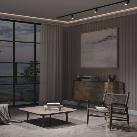 A modern, dimly lit living room has a wooden chair, coffee table, and sideboard beneath a large abstract painting on the striped wall. Floor-to-ceiling windows offer an outdoor view with a plant in the corner, illuminated by Focus LED Single Phase Unity Track Spotlight casting 4000K neutral white light.