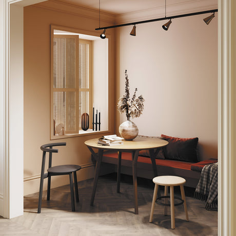 A cozy dining nook features a round wooden table, two chairs, and a cushioned bench. A vase of dried flowers sits atop. The rooms warm beige palette is complemented by a window with decorative screen panels and illuminated by the Virar Single Phase Unity Track Spotlight in matte gold with an adjustable swivel mechanism.