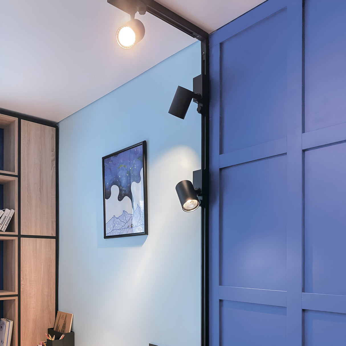 A modern living space with light blue walls, a blue panel door, and wooden shelves features the Vuoro 3-Circuit Trinity Track LED Spotlight 10W 3000K - Black for energy-efficient ceiling lighting. A framed abstract art piece adorns the wall, showcasing minimalist and contemporary design.