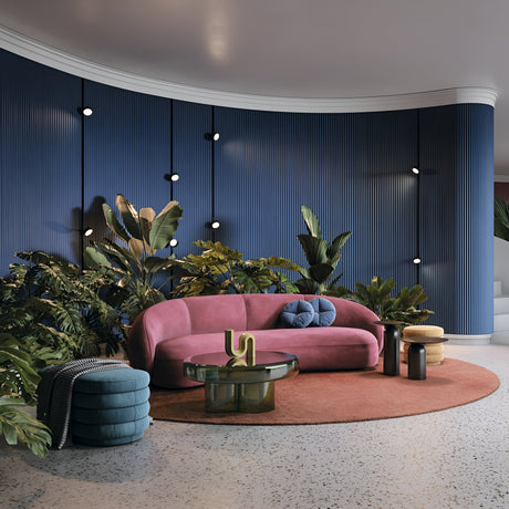 A modern living room includes a pink curved sofa with pillows on a circular brown rug, surrounded by a green glass coffee table and cylindrical side table. The blue textured wall features plants, round lights, and the Rado CCT LED Dimmable Magnetic Exility Track Spotlight for adjustable ambiance.