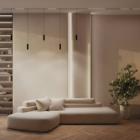 A minimalist living room features beige walls and a modern, modular sofa. The Focus LED Exility Magnetic Track Pendant Light 12W 3000K in black hangs from the ceiling. A large potted plant rests on the right, shelving with decorative items is on the left, and the floor displays a herringbone pattern.