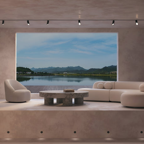 In a modern living room with minimalist furniture, a large window reveals a tranquil lake and distant mountains under a blue sky. The Lipari Single Phase Unity Track Spotlight - Black highlights the round marble table and two cushy white chairs with its adjustable heads.