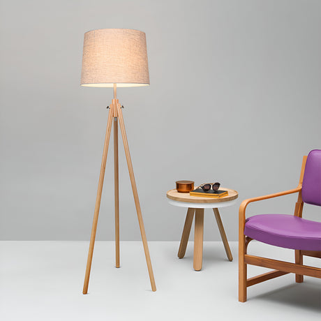A modern living room showcases the Calvin Floor Lamp - Beige with Natural Wood Tripod and minimalist design. Nearby, a small round table holds sunglasses and a cup, paired with a wooden chair featuring purple cushions against a light gray wall.