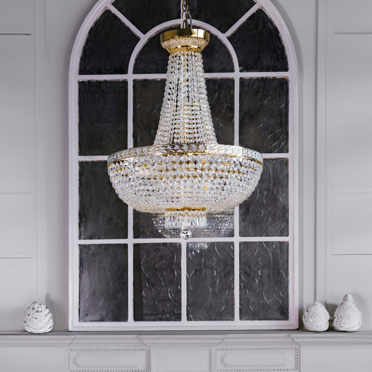 A Bella 8 Light Crystal Chandelier - Gold hangs against an arched window with dark glass panes. The room has white walls and a decorative white shelf adorned with two small white sculptures.