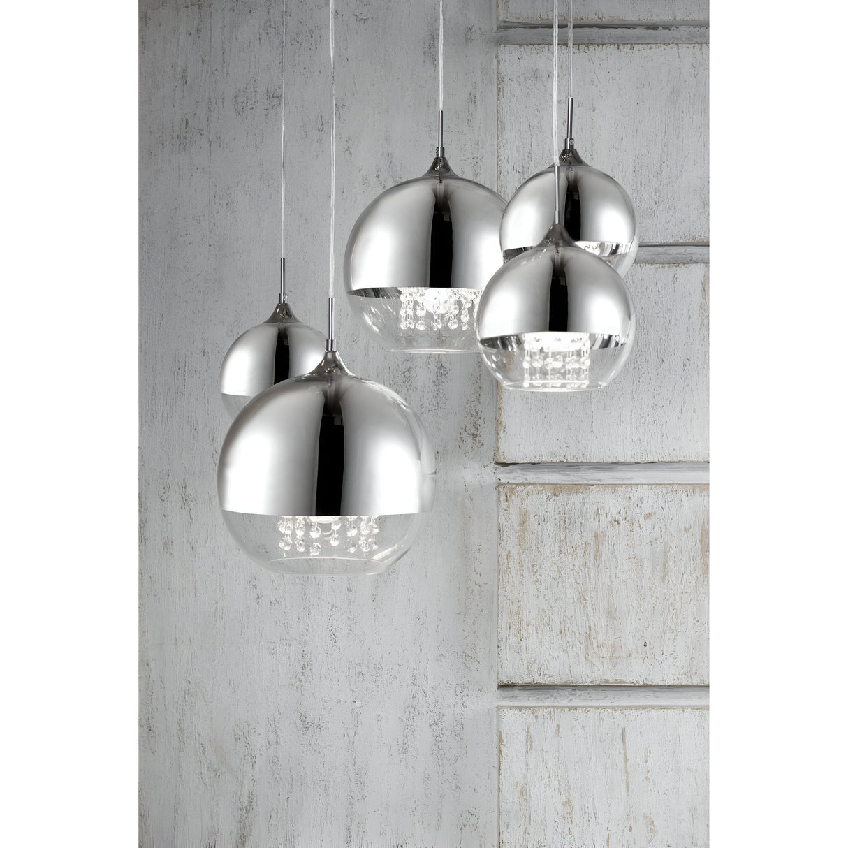 The Fermi 5 Light Cluster Pendant - Nickel features four spherical nickel lamps hanging at varying heights against a textured white wall with vertical lines, offering a shiny, reflective surface that adds a modern, sleek aesthetic to the space.