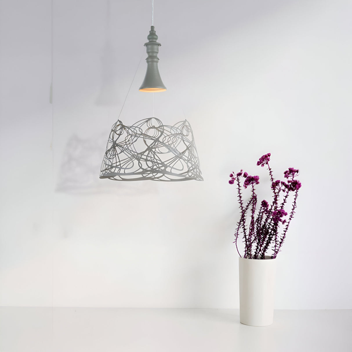 In a minimalist interior, the Elva Single Pendant Light - Grey creates intricate shadows with its patterned fixture and adjustable cord. A white vase with tall purple flowers rests on a white surface against the light gray wall in the background.