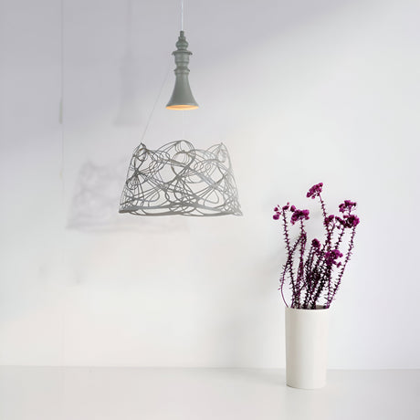 In a minimalist interior, the Elva Single Pendant Light - Grey creates intricate shadows with its patterned fixture and adjustable cord. A white vase with tall purple flowers rests on a white surface against the light gray wall in the background.