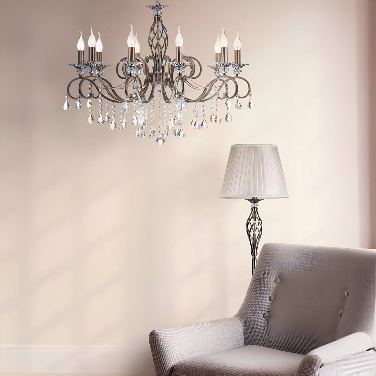 The Grace 10 Light Chandelier in bronze, with candle-shaped bulbs and crystal pendants, adorns the ceiling. Below it, a light gray upholstered armchair sits near a tall floor lamp with a pleated shade against a soft beige wall.