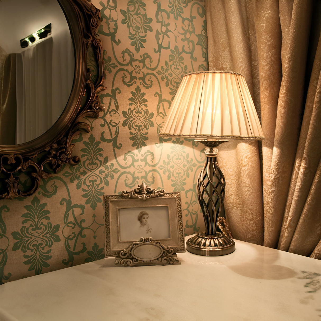 A vintage-style room features a decorative round mirror, the Grace Table Lamp - Bronze with Pleated White Organza Shade, and a framed portrait. The walls display a green and beige damask pattern, complemented by elegant gold curtains.
