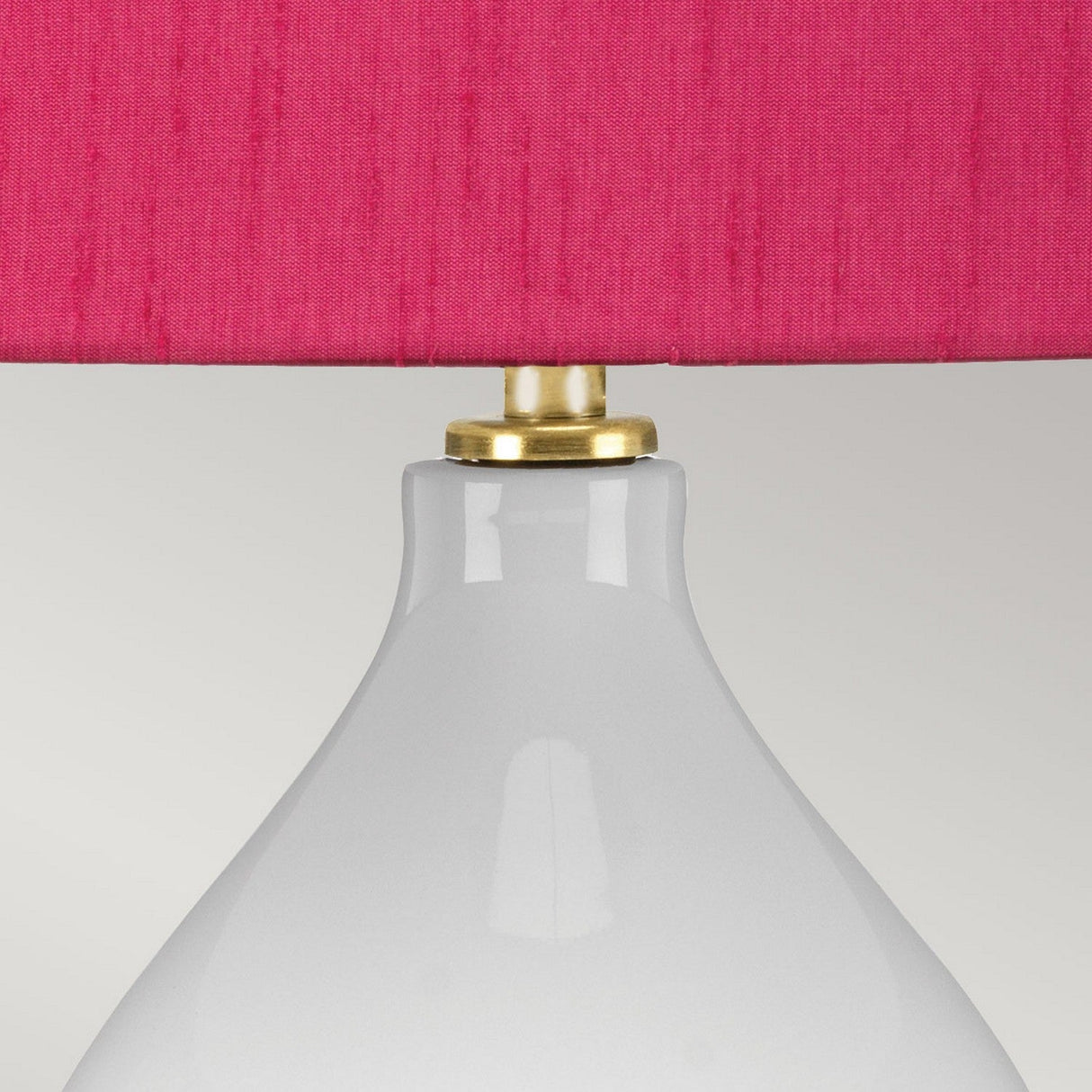 The Isla 1 Light Table Light showcases a detailed view of its sleek white ceramic base paired with a lively pink fabric lampshade. A brass accent elegantly highlights the junction between the base and shade, all set against a subtle light gray backdrop.