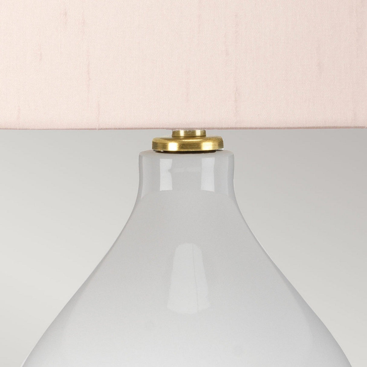 A close-up of the Isla 1 Light Table Light reveals its modern design, featuring a white ceramic base and a light pink fabric lampshade. Brass detailing at the top adds an elegant touch to its sleek appearance.