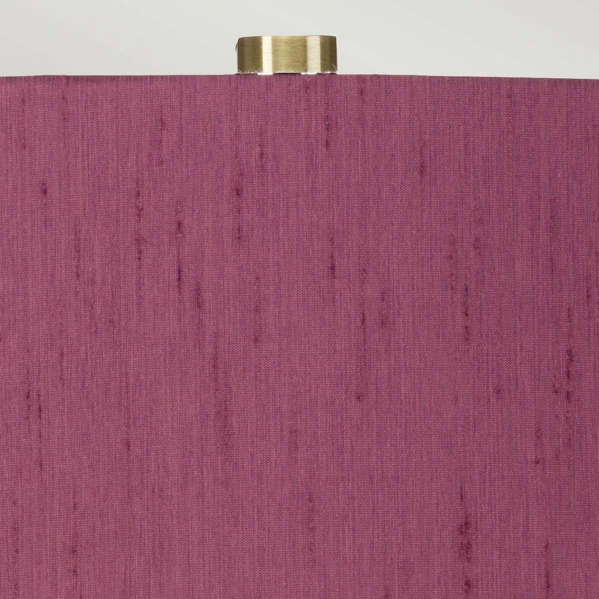 The Isla 1 Light Table Light in white and purple features a textured purple shade with a brass-colored finial on top. The fabric's subtle vertical lines add depth to the color, beautifully contrasting with the ceramic base. A neutral off-white background enhances the elegance of this piece.