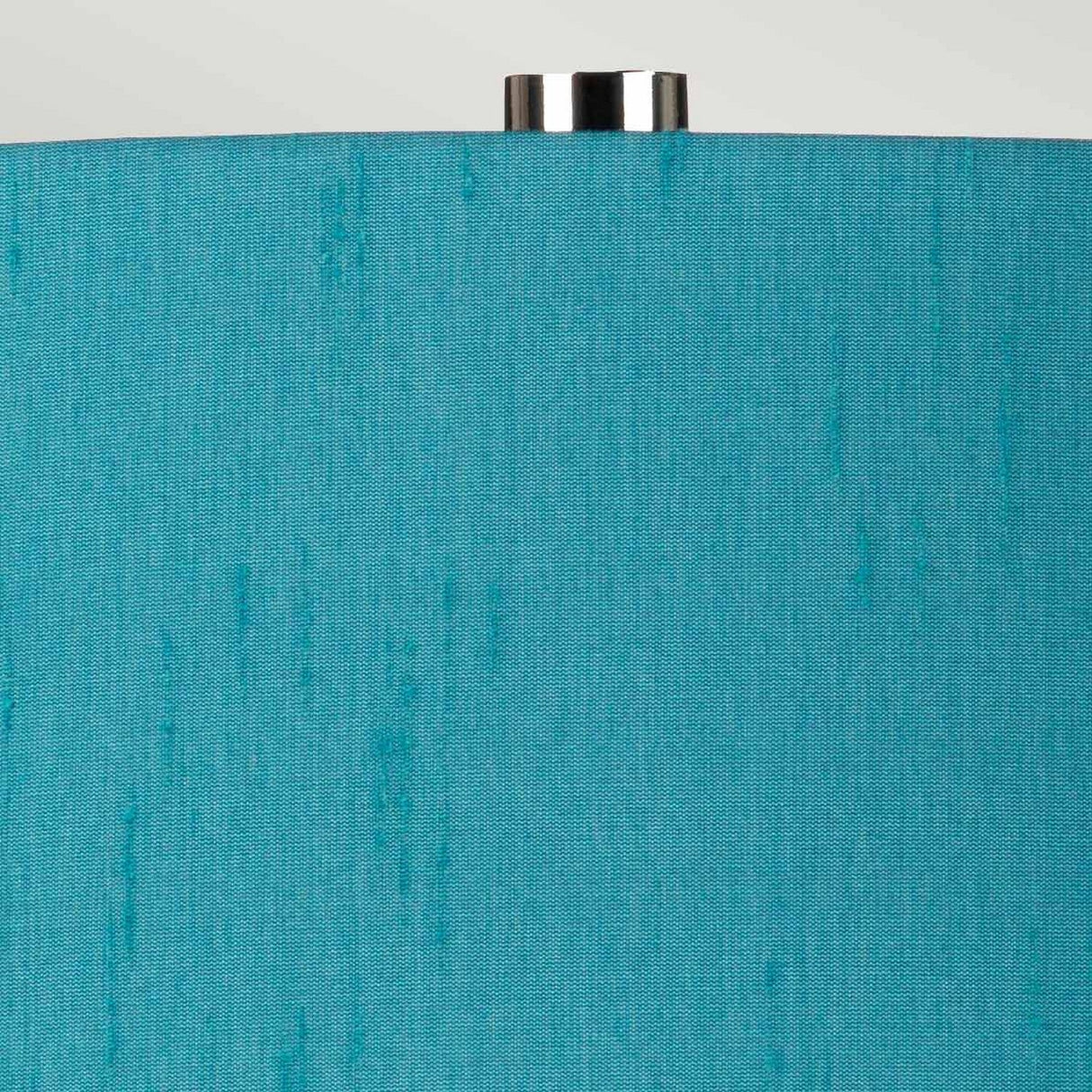 The Isla 1 Light Table Light in White/Teal features a close-up of its elegant teal shade with a subtle textured pattern, accented by a small metallic finial at the top that perfectly complements its ceramic base. Set against a neutral light background, the table light's soothing hue is beautifully highlighted.